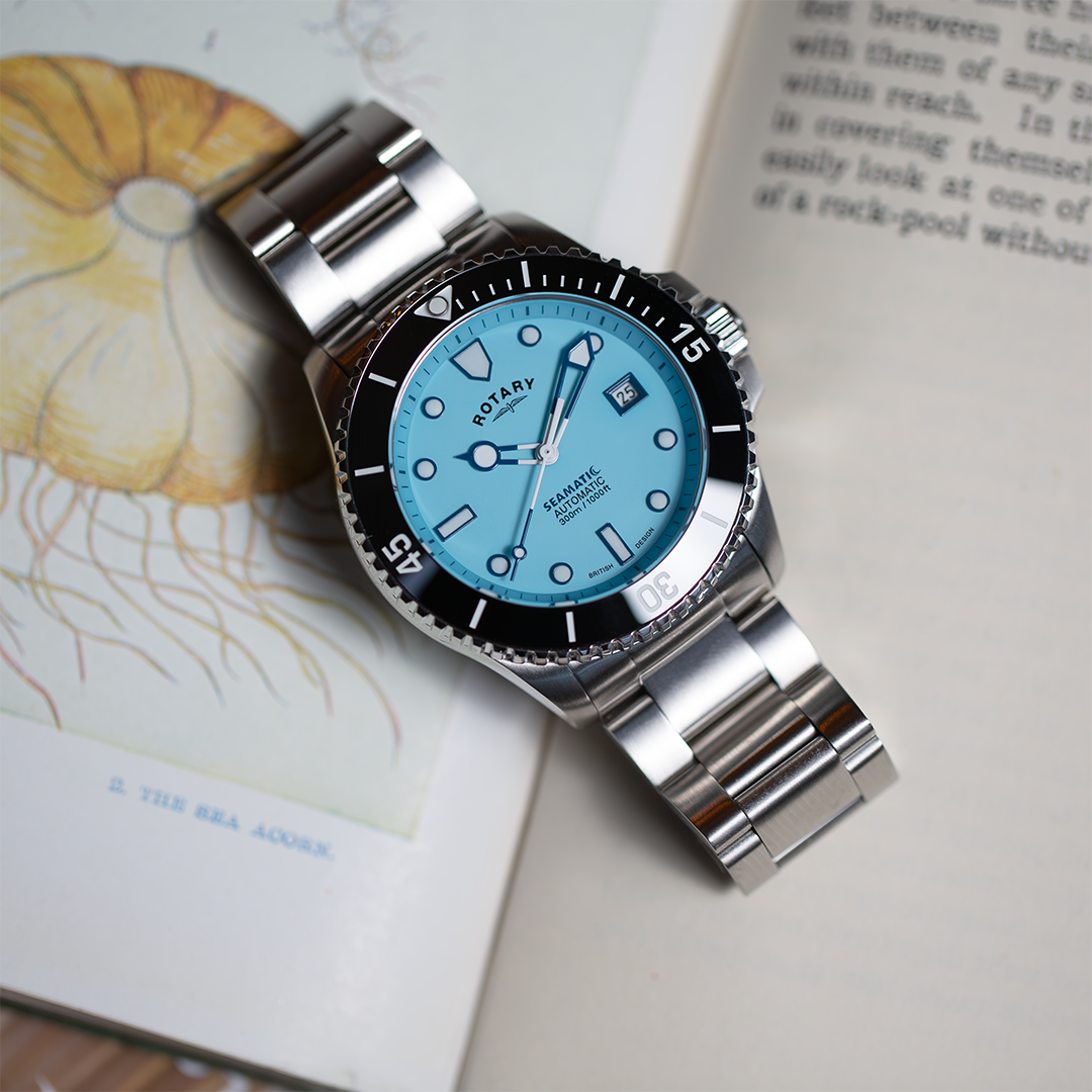 A Rotary Seamatic watch in stainless steel with a baby blue dial. The watch is sat on a vintage nautical themed book