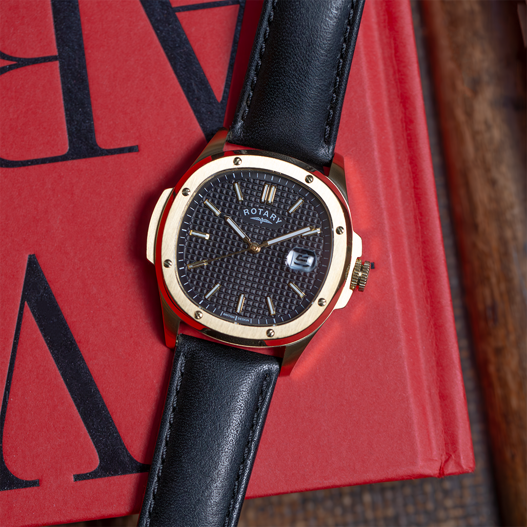 Rotary sport watch in gold with a black leather strap. The watch has a black waffle style dial and a date window at 3. The watch is pictured on a red book on a brown table.